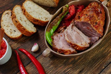 Çatal, bıçak ve rustik koyu arka plan üzerinde ev yapımı rustik ekmek dilimlenmiş domuz loin seramik formundaki pişmiş. Kavrulmuş domuz eti ızgara sebzeler ve domates sosu ile servis. Rahat köyü akşam yemeği