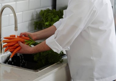 Cook in a white uniform stands on a modern kitchen in a restaurant, washing vegetable clipart