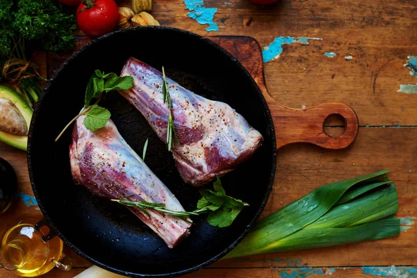 Tuz, karabiber, biberiye ve rustik ahşap masaya kavurma için hazır bir demir tencerede Marine nane ile ham Kuzu İnciği. Yemek hazırlanması. Bir kesme tahtası üzerinde sebze ve otlar. Üstten görünüm.