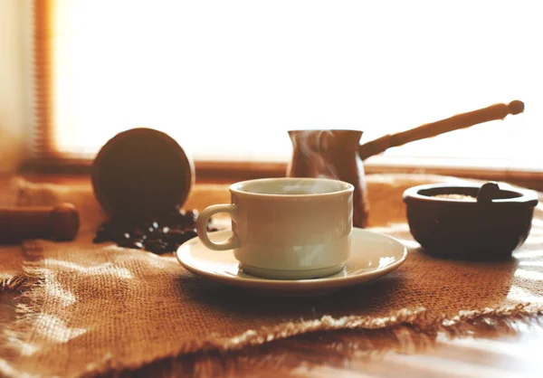 Light falls through the brown shutters on the morning breakfast served coffee in a white small cup, standing next to a cezve which was cooked coffee and brown sugar