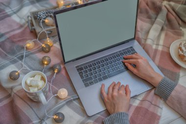 Dizüstü bilgisayarda yazı yazan bir kadın eli. İnternette çalış ve çalış. Sıcak pembe kazak, kış, ev konforu ve rahatlama. Dizüstü bilgisayarlı çalışma alanı, kız eli, pasta, sıcak ekoseli Noel hediyeleri.