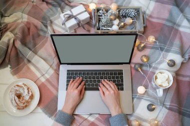 Dizüstü bilgisayarda yazı yazan bir kadın eli. İnternette çalış ve çalış. Sıcak pembe kazak, kış, ev konforu ve rahatlama. Dizüstü bilgisayarlı çalışma alanı, kız eli, pasta, sıcak ekoseli Noel hediyeleri.