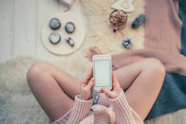 Elinde kahve fincan ıstıracık kazak ve akıllı telefon ile katta kız görüntü. Kısa mesaj veya reklam için kopyalama alanı ile boş ekran. Sıcak rahat ev noel ruh hali. Yumuşak renkler.