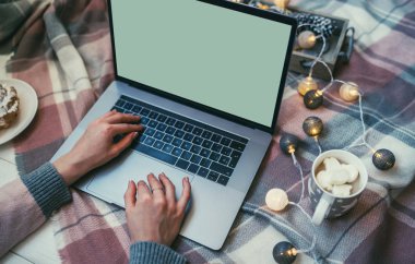 Noel'in tadını evde geçiriyorum. Evde bir katta otururken dizüstü bilgisayar üzerinde çalışan ve kahve içen genç kadın. Boş beyaz ekranlı dizüstü bilgisayarda kullanan ve yazan kızın üst görünümü mockup görüntüsü.