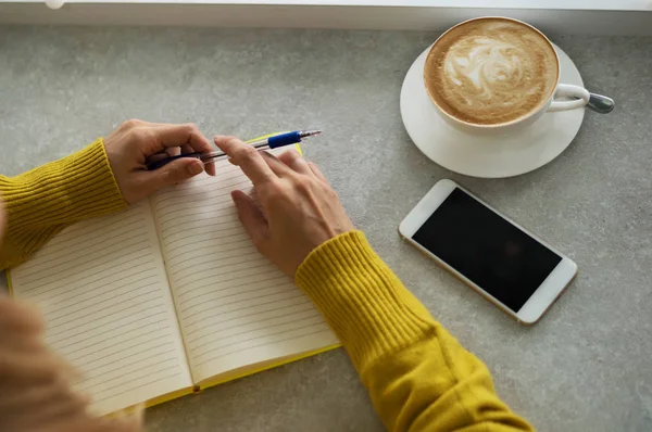 Concept of drinks and youth lifestyle, woman hands and cup of coffee. Young woman writing to do list of goals writing in diary in coffee shop. Female student taking notes in personal organizer.