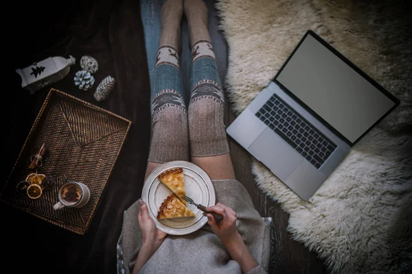 Woman eating cake at Christmas eve. Girl holding plate of christmas cake relaxing at home, drinking cacao, using laptop. Winter and Christmas holidays concept. Top view. Soft, comfy lifestyle.