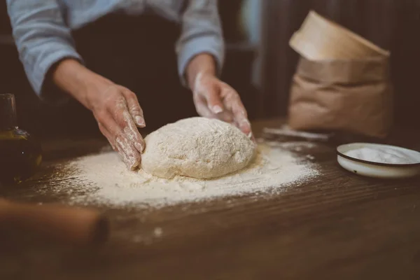 Female hands with dough in ball shape. Basic homemade dough with ingredients on wooden table with natural light. Home healthy food. Low key shot, close up on hands, some ingredients around on table.