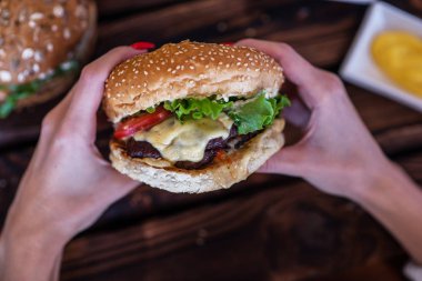 Woman holds burger in hands and potato fries on background in cafe. Hamburger made of beef, onion, tomato, lettuce, cheese and spices. Fresh burger on wooden rustic table.