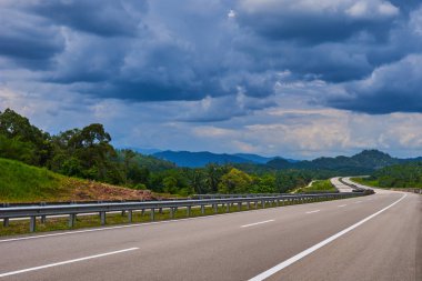 Empty highway or freeway leading to the Cameron Highlands, Malaysia. Spectacular view of the hilly terrain in a blue haze with a cloudy sky and lush tropical rainforest. clipart