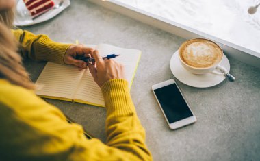 Not defterine yazı yazma üzerine kafa yoran bir kadın. Kafe içinde serbest çalışmaktan hoşlanan genç bir kadın not defterindeki fikirleri not alıyor. Tonlanmış görüntü. Seçici odak.