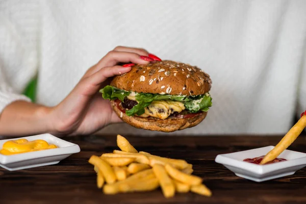 Woman holds burger in hands and potato fries on background in cafe. Hamburger made of beef, onion, tomato, lettuce, cheese and spices. Fresh burger on wooden rustic table.