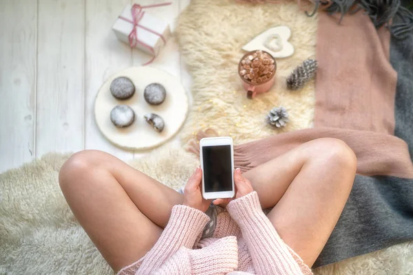 Image of girl in warm sweater on the floor with cup of coffee in hands and smartphone. Blank screen with copy space for your text message or advertising. Warm cozy home christmas mood. Soft colors.