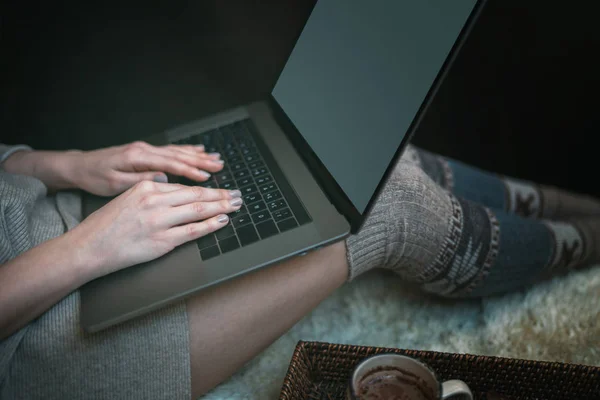 The girl sitting on a wooden floor. Laptop and coffee cup. Feet in woollen socks. The woman relaxes in cozy home. Winter and Christmas holidays concept. Selective focus. Toned image.