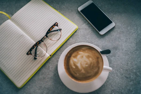 Close up view of freelancer workspace, cup of coffee, paper notepad, smartphone, pen and glasses on table in morning light in the coffee shop interior. Female designer sketching at blank notepad.