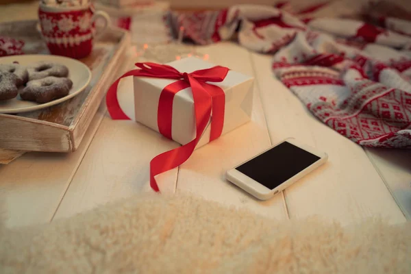 Christmas background with decorations and gift box on wooden floor. Christmas gifts with red ribbon, garland, knitted blanket, smartphone, cup of coffee and cake on white wooden background. Soft focus