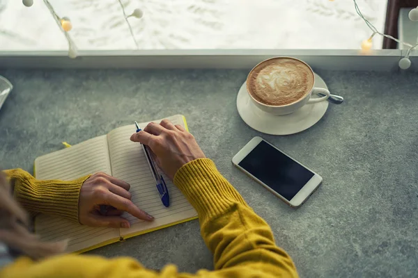 Woman\'s hands writing plans in notes while having a coffee break at the coffee shop table. Woman hand with pencil writing on notebook at coffee shop. Woman working in outdoor at coffee shop.