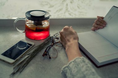 Su ısıtıcısı meyve çayı, kağıt defter, akıllı telefon ve gözlük taş masada kış penceresi önünde. Sabah planlaması ve meyve çayı ile ilham. Kış sıcak içeceği. Tonlu görüntü.