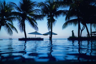 Okyanusa yakın palmiye ağaçlarıyla yüzme havuzu. Şemsiyeler, otel ve tatil tatil açık yüzme merkezi mavi gökyüzünde gün batımı sırasında. Tropik bölgelerde tatiller. Lüks tatil.