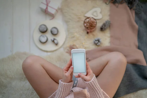 Image of girl in warm sweater on the floor with cup of coffee in hands and smartphone. Blank screen with copy space for your text message or advertising. Warm cozy home christmas mood. Soft colors.