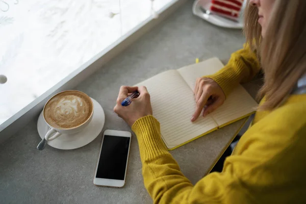 Concept of drinks and youth lifestyle, woman hands and cup of coffee. Young woman writing to do list of goals writing in diary in coffee shop. Female student taking notes in personal organizer.