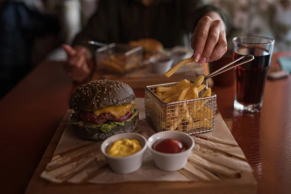 Tasty burger and sauce on wooden tray. Woman eating burger and chips in cafe. People and eating concept. Hamburger, french fries, ketchup, mustard on a wooden board. Toned image.