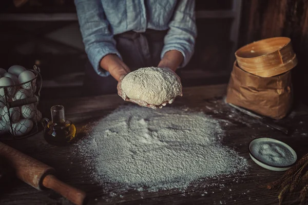 Female hands with dough in ball shape. Basic homemade dough with ingredients on wooden table with natural light. Home healthy food. Low key shot, close up on hands, some ingredients around on table.