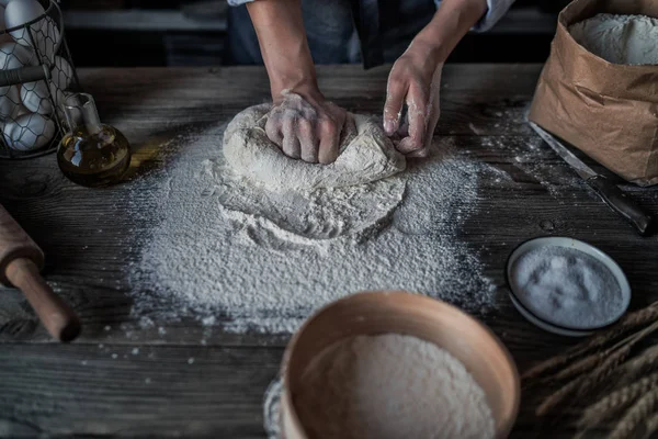 Kneading bread dough for pasta or pizza. Making dough by female hands at bakery. Fresh raw dough for pizza or bread baking on wooden rustic table on warm dark background. Soft focus. Toned image.