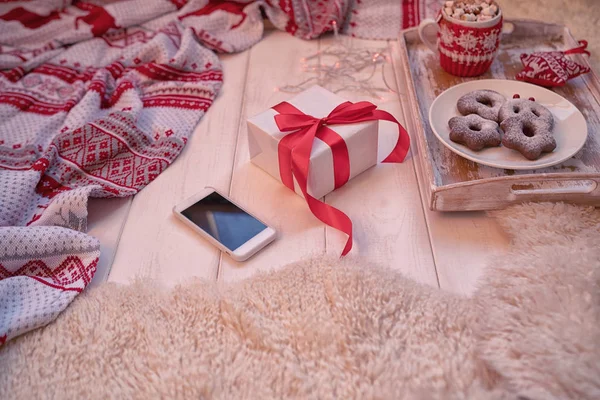 Christmas background with decorations and gift box on wooden floor. Christmas gifts with red ribbon, garland, knitted blanket, smartphone, cup of coffee and cake on white wooden background. Soft focus