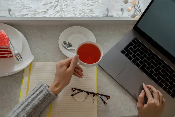 Entrepreneur with a laptop relaxing in a coffee shop enjoying a cup of fruit tea. Hot tea cup on a frosty winter window background. Cozy coffee shop atmosphere in winter. Selective focus.