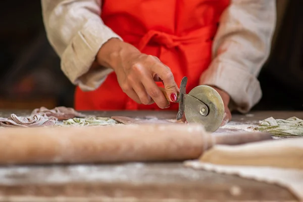 Senior woman cook making tagliatelle pasta with dough in restaurant kitchen. Making pasta. Making pasta in restaurant. Colored raw vegetable pasta with beets, carrots and spinach.