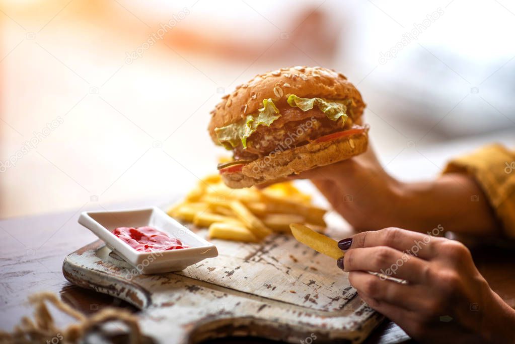 Young woman is eating fast food. Woman is eating burger while spending time in cafe. Tasty vegan burger with lettuce and beet served on cutting board. Side view with free spase for you text.