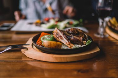 Baharatlı Izgara kuzu bacak sebze ve otlar üzerinde bir kavurma ile dökme demir. Sebzeli ızgara kuzu. Sağlıklı gıda. Yemek ve eğlence kavramı. Kadın gıda ile masada yemek yiyorlar. Tonlu görüntü.