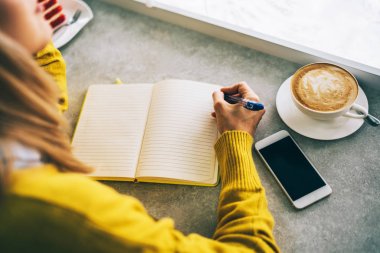 Not defterinde yayın yazma oluşturma üzerinde kafa yoran pensive kadın yazma. Kafe iç mekanda serbest çalışma yapmaktan zevk alan genç kadın not defterinde fikir notları hazırlıyor. Tonlu görüntü. Seçici odaklama.