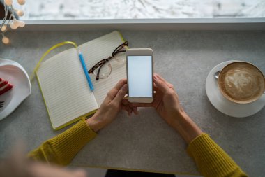 Kahve dükkanında sıcak kahve fincanı ve kek ile boş beyaz ekran ile beyaz cep telefonu tutan ellerin kırpılmış görüntü. Ücretsiz wifi bağlı akıllı telefon gelen sms mesajı okuma Kadın.