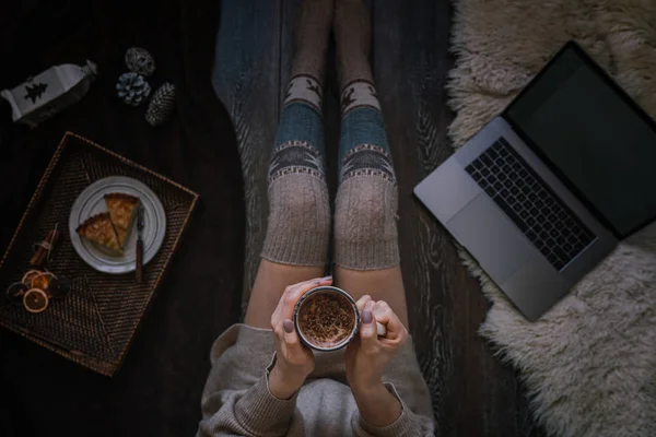 Woman in cozy home wear relaxing at home, drinking cacao, using laptop. Hot chocolate or cocoa with marshmallow and spices. Winter and Christmas holidays concept. Top view. Soft, comfy lifestyle.