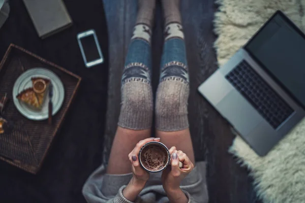 Woman in cozy home wear relaxing at home, drinking cacao, using laptop. Hot chocolate or cocoa with marshmallow and spices. Winter and Christmas holidays concept. Top view. Soft, comfy lifestyle.