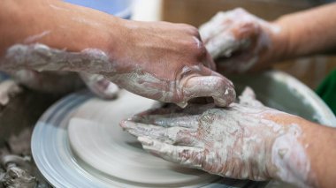 Islak çamura bulanmış ellerin yakın çekimi, iki kişi dönen tekerleğin üzerinde bir çömlek parçasını şekillendirirken, seramik işçilikte yer alan takım çalışmasını ve sanatını vurguluyor..