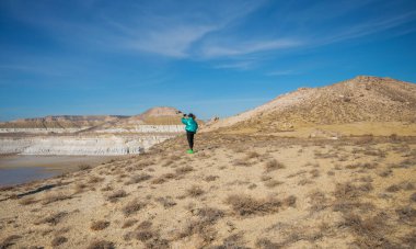 Turkuaz ceketli bir gezgin, kurak düzlüklerde beyaz kayalıklarla ve parlak mavi gökyüzünün altında katmanlı kaya oluşumlarıyla selfie çeker..