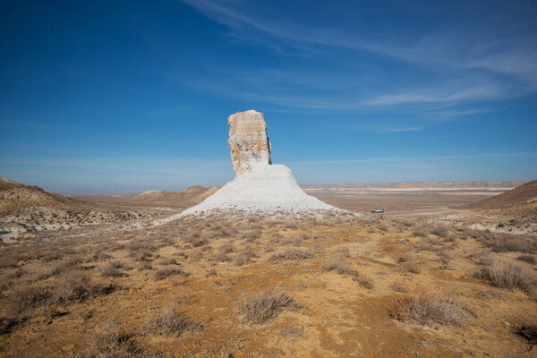 A striking white limestone rock formation rises in a vast arid steppe under a bright blue sky. A vehicle in the distance adds scale to the remote landscape.
