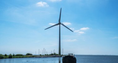 Yükselen rüzgar türbini, limanlı bir teknesi ve yemyeşil denizaltılarıyla çevrili ve sürdürülebilir yeniliğin güzelliğini yansıtan uzak yelkenlileri olan sakin bir sahil kenarında duruyor..