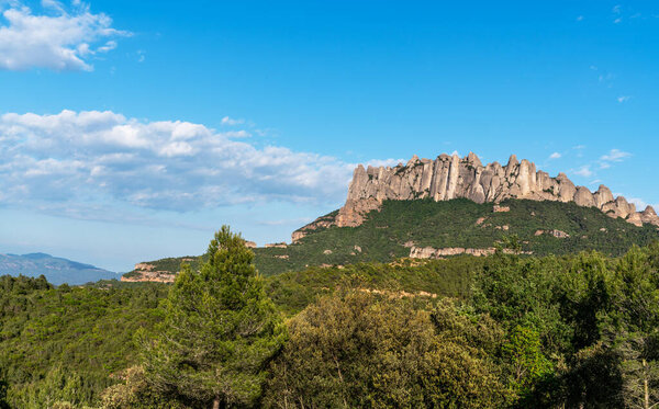 A breathtaking view of the rugged Montserrat mountain range with its distinctive rock formations, lush green forests, and clear blue sky, showcasing the natural beauty of the Catalonian landscape.