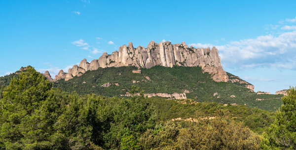 A breathtaking view of the rugged Montserrat mountain range with its distinctive rock formations, lush green forests, and clear blue sky, showcasing the natural beauty of the Catalonian landscape.