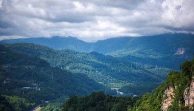 Ağaçlarla kaplı bir uçurumun çarpıcı bir görüntüsü. Kayaların farklı katmanlı oluşumları, kıvrımlı bir yolu olan yemyeşil bir vadiye bakıyor. Puslu dağların ve dinamik bir gökyüzünün zeminine kurulmuş.