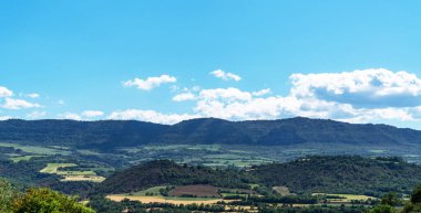 Parlak mavi gökyüzünün altındaki İspanyol kırsalının kayan tepelerini ve yamalı arazilerini manzarası. Yeşil ve kahverenginin tonları, kabarık beyaz bulutlarla çok güzel tezat oluşturuyor.