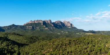 İspanya 'nın Katalonya kentindeki Montserrat dağlarının berrak mavi gökyüzünün altında çarpıcı bir manzarası. Pürüzlü zirveler yemyeşil çayırların üzerinde dramatik bir şekilde yükselir..