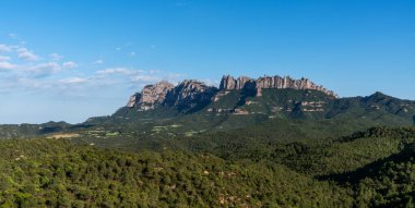 İspanya 'nın Katalonya kentindeki Montserrat dağlarının berrak mavi gökyüzünün altında çarpıcı bir manzarası. Pürüzlü zirveler yemyeşil çayırların üzerinde dramatik bir şekilde yükselir..