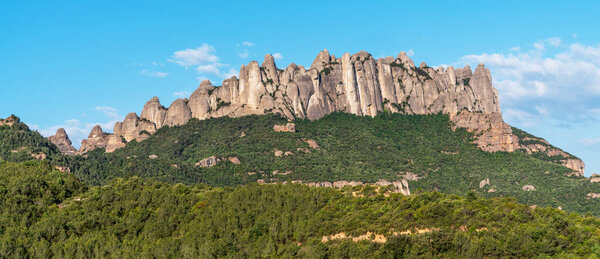A breathtaking view of the rugged Montserrat mountain range with its distinctive rock formations, lush green forests, and clear blue sky, showcasing the natural beauty of the Catalonian landscape.
