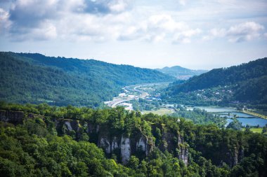 Parlak yeşil tepeleri kesen kıvrımlı bir nehri olan pitoresk bir manzara paralel uzanan bir otoyol ve dağınık yerleşim alanları, hepsi parçalı bulutlu bir gökyüzünün altında.