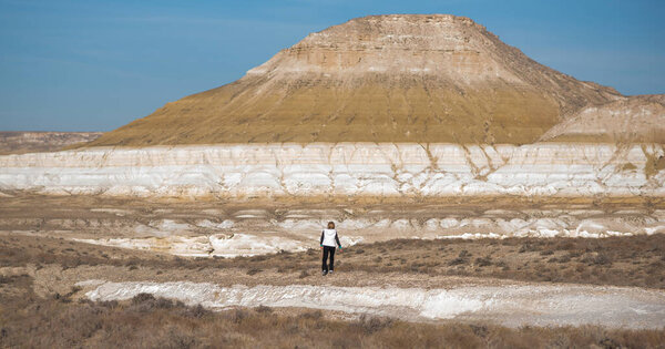 A person walking in a dry desert landscape with a prominent yellow and white layered mountain in the background under a clear blue sky, showcasing a unique geological formation.