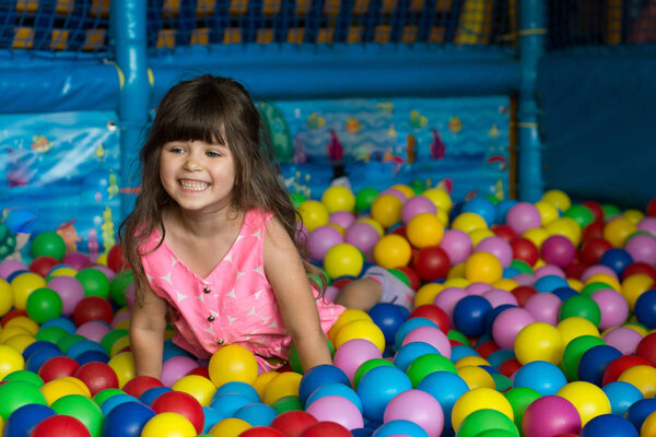 Happy laughing girl having fun in ball pit at indoor play center. Child playing with colorful balls in playground ball pool.
